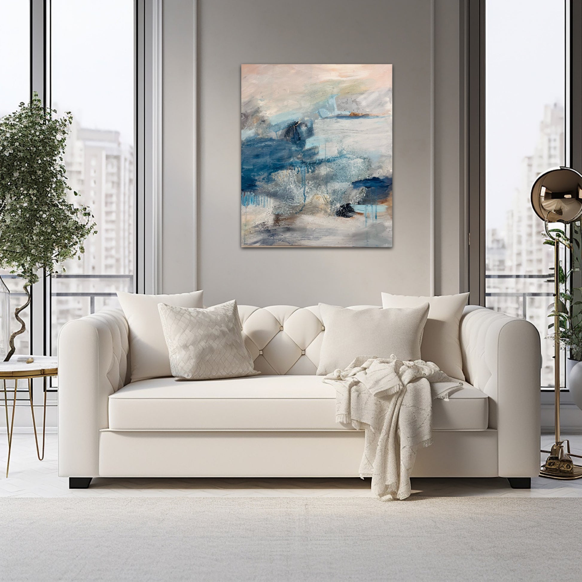 A lifestyle photo of an atmospheric landscape painting in blues, peach and grey, above a cream couch on neutral colour  walls