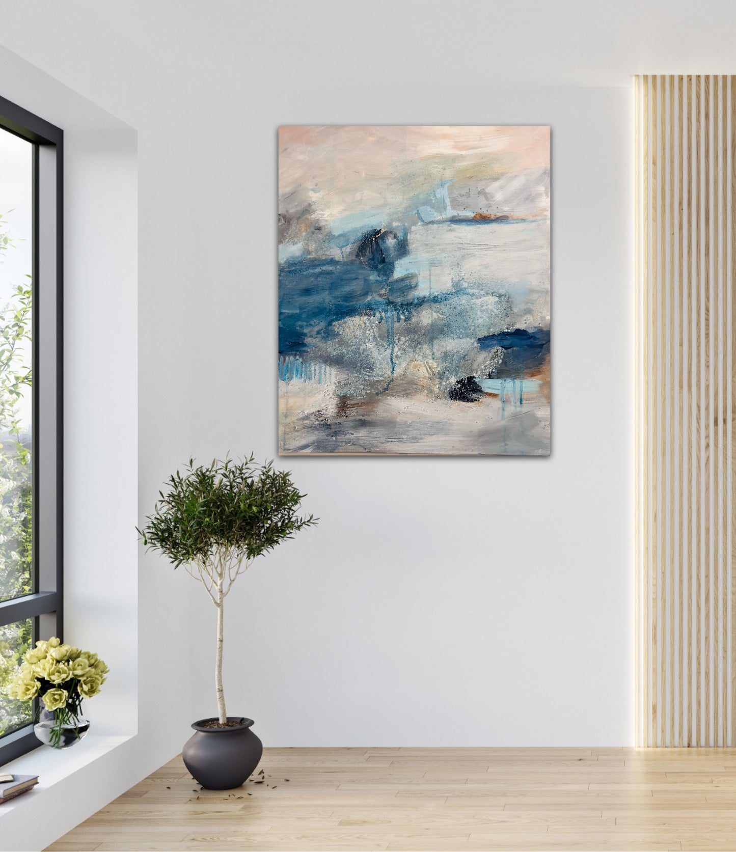 Atmospheric painting in blues and peach hung on a white wall next to an olive tree in a plant pot with light shining in from the window to the side