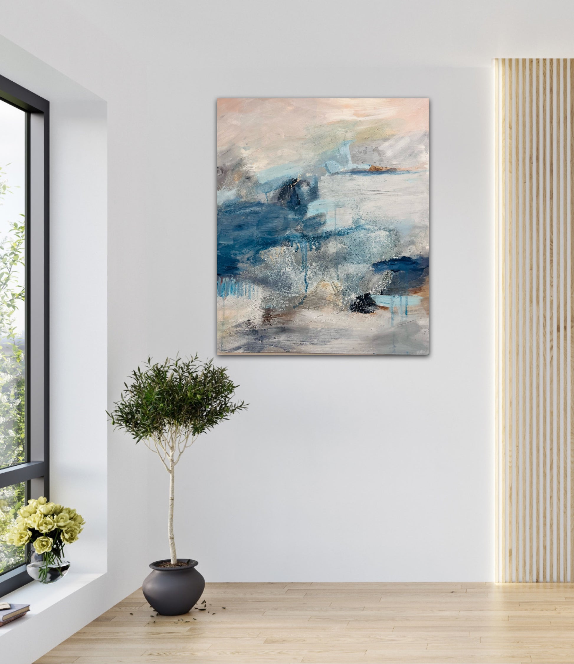Atmospheric painting in blues and peach hung on a white wall next to an olive tree in a plant pot with light shining in from the window to the side