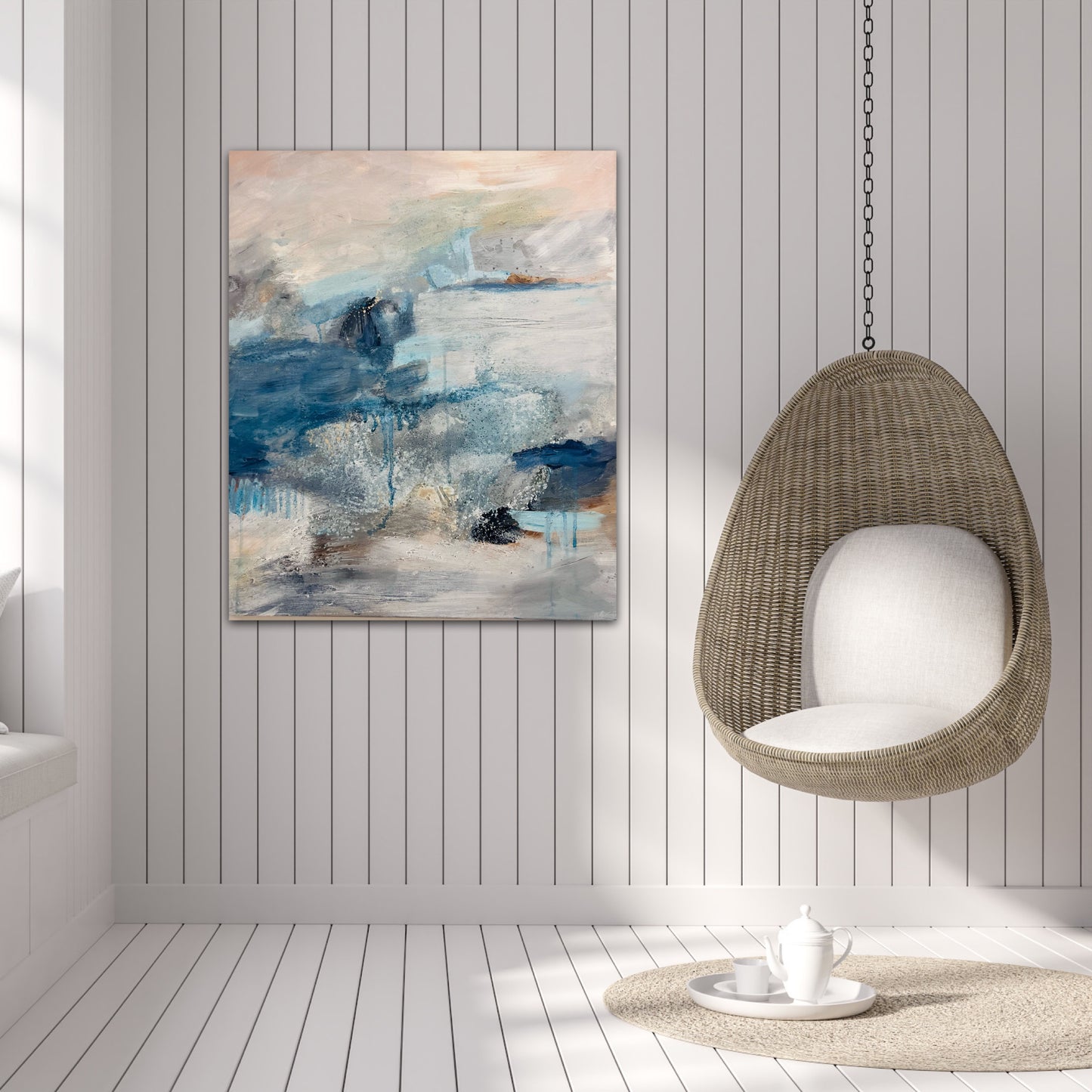 Abstract landscape painting in peach, blue and grey hung on a white panelled wall next to a wicker hanging egg chair