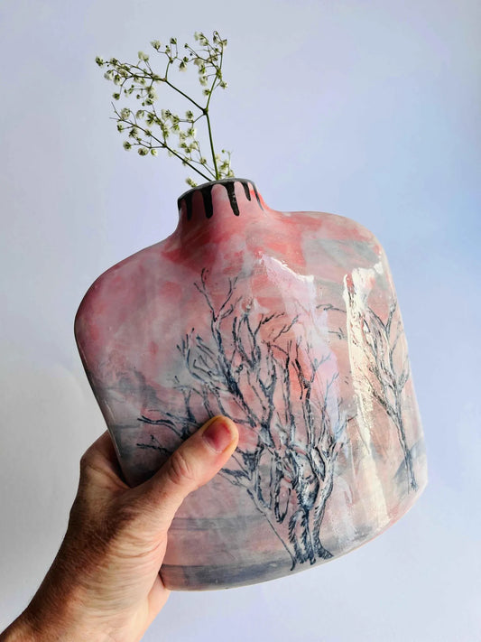 Hand holding a pink ceramic vase with tree design against a light blue background