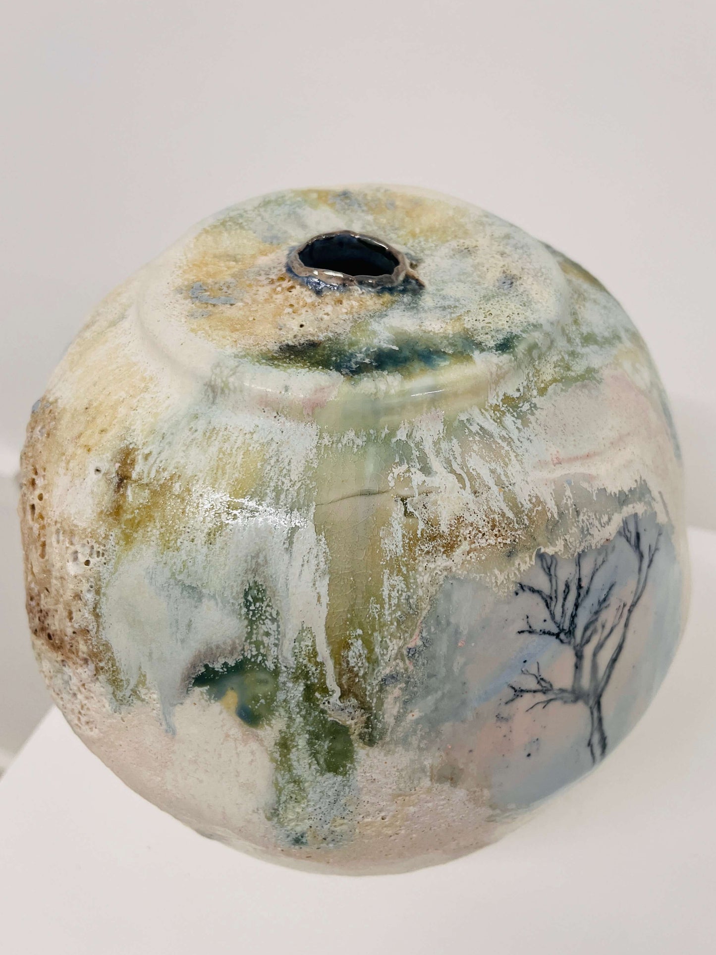 A handmade ceramic vase with a rounded, bulbous shape and a small hole at the top for pouring or hanging. The vase features a textured, mottled glaze with cream, beige, light blue, green, and brown tones, along with a dark blue painted tree design on the side, set against a plain white background.