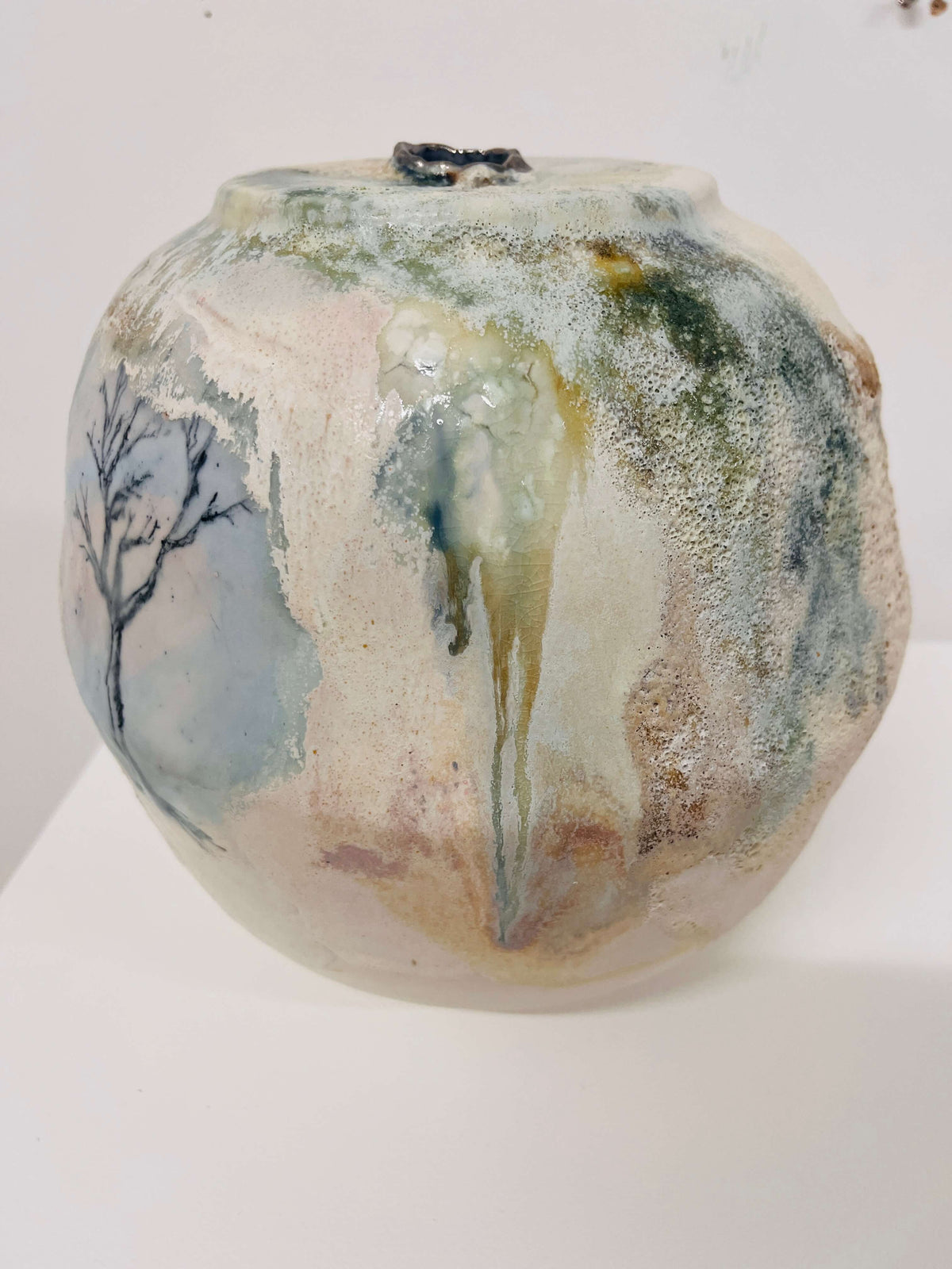 A handmade ceramic pot with a rounded shape, featuring a textured, glossy glaze with drips of green, blue, and beige over a cream base, decorated with a black painted tree on the left side.