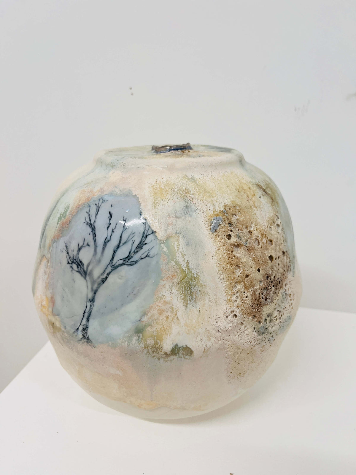 A handmade ceramic pot with a textured, organic surface featuring a painted tree design on a light blue-gray background, alongside earthy brown and beige mottled patterns, set against a plain white background.
