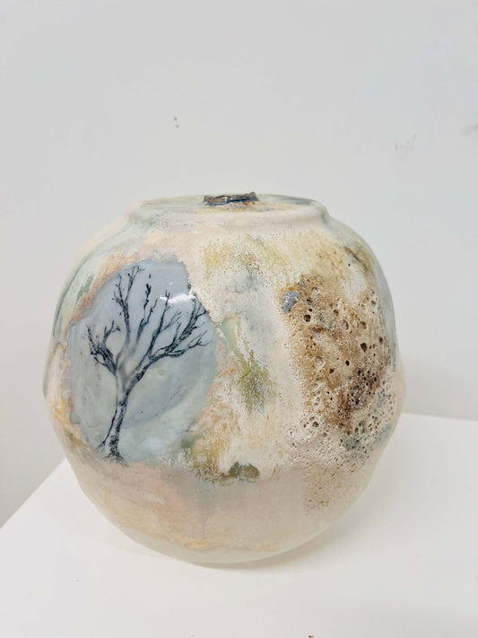 A handmade ceramic pot with a textured, organic surface featuring a painted tree design on a light blue-gray background, alongside earthy brown and beige mottled patterns, set against a plain white background.